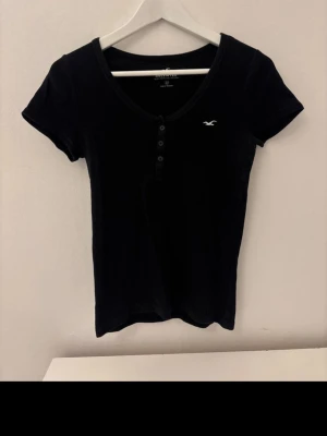 Svart ribbad t-shirt från Hollister - Svart t-shirt från Hollister med ribbad struktur och v-ringning. T-shirten har korta ärmar, knappar framtill och en liten vit logga på bröstet. Snygg och enkel modell som sitter tight och har en klassisk look. Storlek M men passar XS
