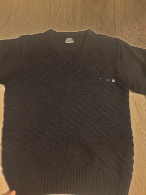 Svart stickad tröja med v-ringning - Svart stickad tröja från Şahin Tekstil med v-ringad hals och snyggt diagonalt mönster. Tröjan har långa ärmar och coola broderade X-patchar på armbågarna. Ribbstickade muddar vid ärmslut och nederkant ger en schysst passform.