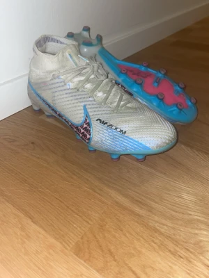 Nike Mercurial Air Zoom fotbollsskor - Säljer ett par Nike Mercurial Air Zoom Elite fotbollsskor i vitt med blå och rosa detaljer. Skorna har snörning, hög strumpa och mönstrad swoosh i svart och rosa. Sulan är blå med rosa dobbar och AIR ZOOM-tryck. Perfekt för dig som vill sticka ut på planen. Ej använda länge eftersom jag slutat på fotboll. Jag skulle klassa dom på ett stabilt 8,5/10 i skick. Nypris: Ca 3000! Pris kan diskuteras vid snabbt köp