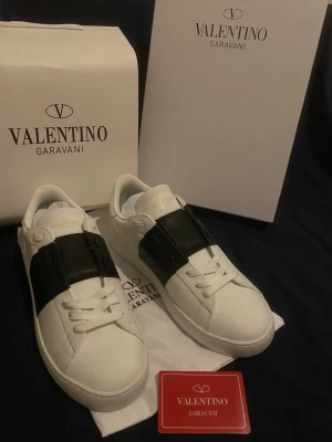 Valentino Garavani Open sneakers - Säljer ett par snygga Valentino Garavani Open sneakers i vitt skinn med bred svart läderdetalj över mitten. Klassisk rund tå, platt sula och vita snören. Märkeslogga på plösen och medföljer originalkartong och dustbag.