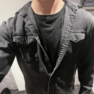 Svart jeansskjorta med knappar - Snygg svart jeansskjorta med knappar och två bröstfickor. Perfekt för en cool och avslappnad stil. Skjortan har långa ärmar och en klassisk krage.