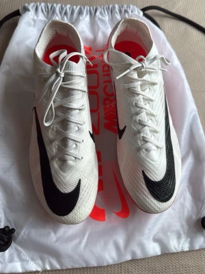 Nike Mercurial Zoom fotbollsskor 40.5 - Säljer ett par Nike Mercurial Zoom fotbollsskor i vitt med svarta och röda detaljer. Skorna har snörning, platt sula med dobbar och är tillverkade i ett lätt syntetmaterial. Perfekta för dig som vill ha grym känsla på planen och sticker ut med snygg design.