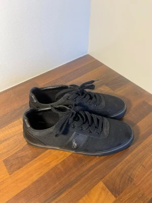 Svarta sneakers från Polo Ralph Lauren - Säljer ett par svarta sneakers från Polo Ralph Lauren med klassisk logga på sidan och POLO-tryck på plösen och hälen. Skorna har snörning, rund tå och är tillverkade i textil med detaljer i skinnimitation. Skorna är knappt använda då de inte passade bra. Hör av er vid frågor!