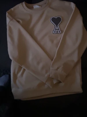 Beige sweatshirt från Ami Paris - Säljer en beige sweatshirt från Ami Paris med det ikoniska hjärta-A-märket broderat på bröstet. Tröjan har rund halsringning och långa ärmar. Tillverkad i mjuk bomull för en skön och avslappnad look.