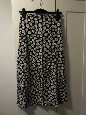 Blommig kjol från DeFacto - Supersöt kjol från DeFacto med svart botten och vita prästkragar över hela tyget. Kjolen har en rak och flowy passform som ger en chill vibe. Perfekt för dig som vill ha en statement piece med blommigt mönster. Materialet känns lätt och skönt. Den har en dragkedja vid sidan.