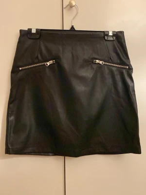 Svart skinnkjol från BikBok - Säljer en svart kort skinnkjol från BikBok med två sneda dragkedjor framtill som cool detalj. Kjolen har en clean look och passar perfekt till en edgy outfit. Materialet är mjukt och följsamt.