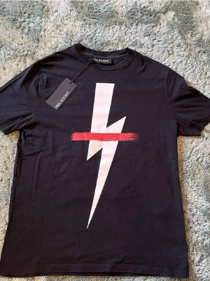 Neil Barrett svart t-shirt  - Svart t-shirt från Neil Barrett med stor vit blixt och röd linje över bröstet. Klassisk rund hals och korta ärmar. Tillverkad i mjuk bomull för skön känsla och snygg passform. Perfekt statement-plagg för dig som gillar grafiska tryck.