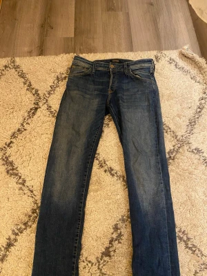 Blå jeansbyxor från Jack & Jones - Snygga blå jeans från Jack & Jones i klassisk rak modell. Jeansen har fem fickor, kontrastsömmar och en brun patch bak i midjan. Perfekta till vardagslooken och passar till det mesta. Storlek 31/34.