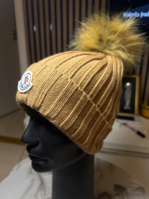 Beige stickad mössa från Moncler - Snygg beige stickad mössa från Moncler med ribbad struktur och stor fluffig pälsboll på toppen. Märkeslogga framtill. Perfekt accessoar för att hålla värmen och se trendig ut under kyliga dagar.