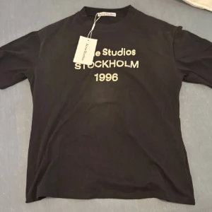 Svart Acne Studios t-shirt Stockholm - Svart t-shirt från Acne Studios med tryck 'Stockholm 1996' i vitt framtill. Klassisk rund hals och korta ärmar. Tillverkad i 100% bomull för en skön och avslappnad känsla. Perfekt för dig som gillar stilrena och trendiga plagg.