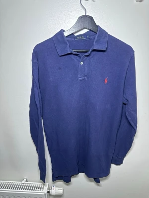 Blå långärmad pikétröja Polo Ralph Lauren - Klassisk marinblå långärmad pikétröja från Polo Ralph Lauren med röd broderad logga på bröstet. Tröjan har krage, knappar vid halsen och är gjord i mjuk bomull. Perfekt till en clean och avslappnad stil.