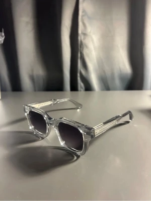 Grå transparenta solglasögon CHIMI - Snygga solglasögon från CHIMI med transparent grå båge och fyrkantig form. Glasen är mörka med en lätt blå ton. Kommer med originalväska och fodral. Perfekt accessoar för att lyfta din stil. Priset är justerbart!