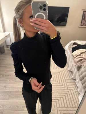 Svart polotopp med puffärmar - Säljer en svart polotopp med långa puffärmar och figurnära passform. Toppen har en hög krage och är enkel men ändå stilren, perfekt att matcha med både jeans och kjol. Materialet känns mjukt och stretchigt.