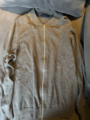 Grå kofta med dragkedja från Berkelly - Snygg grå kofta från Berkelly med hel dragkedja framtill och ribbade muddar vid hals, ärmslut och nederkant. Mjuk och skön stickad känsla, perfekt för lager-på-lager. Enkel design som funkar till många stilar.