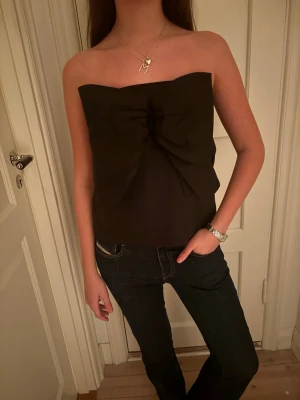 Svart axelbandslös tubtopp med rosett - Säljer en svart axelbandslös tubtopp med stor rosettdetalj framtill. Toppen har en stilren och elegant look, perfekt att styla med jeans eller kjol. Fungerar lika bra att använda som kjol