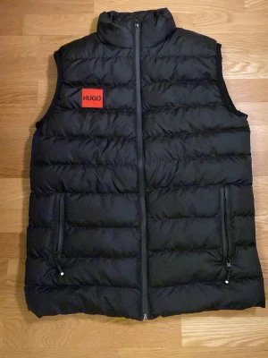 Svart dunväst från Hugo Boss - Säljer en svart dunväst från Hugo Boss med quiltad design och hög krage. Västen har dragkedja framtill, två fickor med dragkedja och en röd Hugo-logga på bröstet. Perfekt för lager-på-lager och snygg till streetwear.
