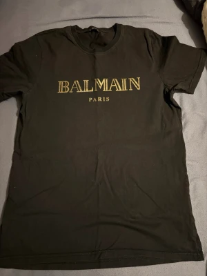 Svart Balmain t-shirt med guldtryck - Svart t-shirt från Balmain med ikoniskt guldigt logotryck på bröstet. Klassisk rund halsringning och korta ärmar. Tillverkad i mjuk bomull som känns skön mot huden. Perfekt för dig som vill ha en clean men ändå lyxig vibe.