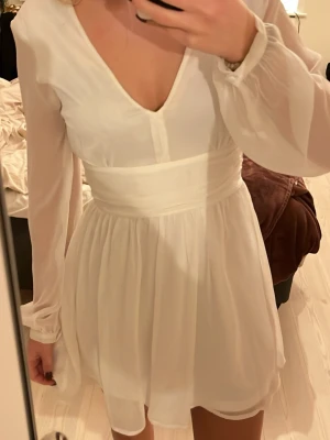 Vit omlottklänning från Bubbleroom - Supersöt vit kortklänning från Bubbleroom med v-ringning, långa transparenta ärmar och omlottdetalj i midjan som knyts baktill. Kjolen är lätt utställd och tyget känns luftigt och flowy. Perfekt för sommaren eller studenten! 