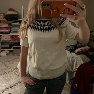 Vit mönstrad tröja Jumperfabriken S - Kortärmad vit tröja från Jumperfabriken i storlek S. Tröjan har ett svart grafiskt mönster runt halsen och ribbade muddar nedtill och vid ärmslut. Tillverkad i mjuk bomull, perfekt för vår och sommar. Snygg och enkel att styla till jeans eller kjol.