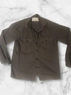 Grå grön overshirt från A days march - Säljer en oliv overshirt med klassisk krage och knäppning framtill. Jackan har två stora bröstfickor med lock och knappar, samt långa ärmar. Perfekt att slänga över en hoodie eller t-shirt för en chill look.