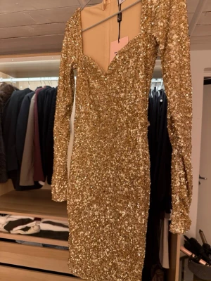 Guldig paljettklänning från Oh polly - Säljer en glittrig guldig kortklänning från Oh polly med långa ärmar och djup v-ringning. Klänningen är täckt av paljetter och har en figurnära passform som verkligen sticker ut. Endast provad, klänningen sticks inte eller är oskön.
