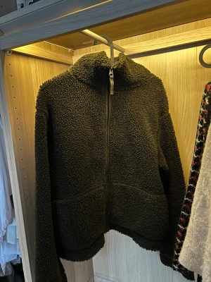 Olivgrön teddyjacka med dragkedja - Mysig mörkbrun teddyjacka med hög krage och hel dragkedja framtill. Jackan har två stora fickor och är tillverkad i ett fluffigt teddy-material som håller dig varm. Perfekt för kyliga dagar och enkel att styla med olika outfits.