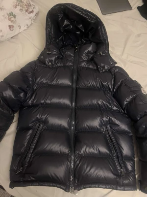 Svart Moncler pufferjacka med huva - Säljer en svart pufferjacka från Moncler med glansig finish och avtagbar huva. Jackan har två stora fickor med dragkedja, Moncler-logga på ärmen och coolt serietryck på insidan. Perfekt för kalla dagar och riktigt snygg streetstil. Priset är inte ristat i sten men eftersom varan är äkta är priset högt.
