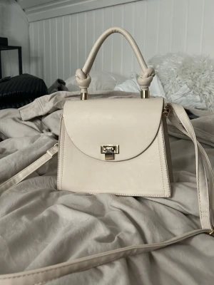 Beige axelväska med gulddetaljer - Snygg beige axelväska i stilren design med guldfärgat spänne och detaljer. Väskan har ett handtag med knutdetaljer och en lång axelrem som går att justera. Perfekt storlek för det viktigaste och ger en clean look till din outfit.