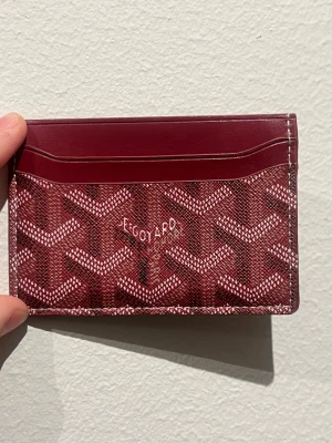 Röd plånbok med mönster - Aldrig använd snygg röd plånbok  med grafiska mönster i vitt och vinrött. Plånboken har flera kortfack och är tillverkad i skinn med detaljer i syntet. Perfekt för dig som vill ha något stilrent och lyxigt i fickan.