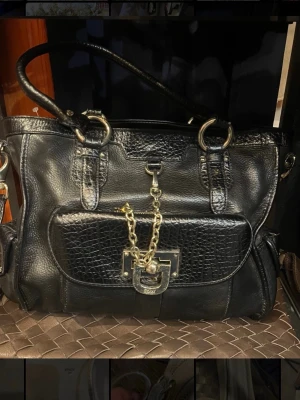 Svart handväska från DKNY i läder - Säljer en svart handväska från DKNY i läder med snygga guldfärgade metalldetaljer och kedja framtill. Väskan har strukturmönstrad frontficka, dubbla handtag och dragkedja upptill. Perfekt för dig som vill ha en stilren och lyxig accessoar. Väskan stängs både med magnet och dragkedja samt har många olika praktiska fack i väskan. Orginalpris på väskan är runt 1500kr.  Väskan säljs i befintligt skick då den har använts! Den har dock använts sparsamt och ansvarsfullt. Hör av dig vid intresse!😊