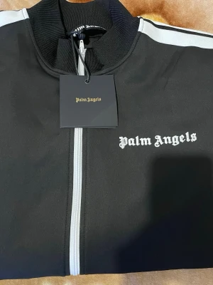 Palm Angels svart track jacket - Säljer en svart track jacket från Palm Angels med vita ränder längs ärmarna och vit dragkedja framtill. Tröjan har hög krage, ribbade muddar och logga broderad på bröstet. Perfekt för en sportig och trendig look.