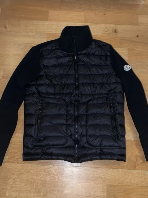 Svart quiltad jacka från Moncler - Säljer en svart quiltad jacka från Moncler med stickade ärmar och hög krage. Jackan har dragkedja framtill, två sidofickor och klassisk Moncler-logga på ärmen. Materialmix med blankt syntet på kroppen och stickad struktur på ärmarna ger en riktigt clean look.
