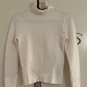 Vit stickad tröja med dekorativa knappar från Zara  - Vit stickad tröja med långa ärmar och dekorativa blomformade knappar vid ärmsluten från Zara i storlek S. Tröjan har en ribbad kant och en hög krage med volangdetalj. Knappt använd. 