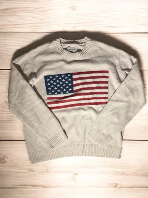 Vit stickad tröja Hollister USA-flagga - Vit stickad tröja från Hollister med stor amerikansk flagga på bröstet. Tröjan har rund halsringning, ribbade muddar och är långärmad. Perfekt för dig som vill ha en klassisk college-look med en twist.