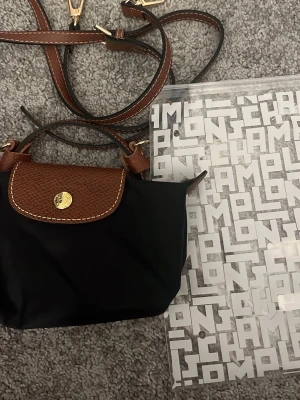 Svart axelväska från Longchamp - Säljer en svart axelväska från Longchamp med bruna skinn-detaljer och guldfärgad knapp. Väskan har justerbar axelrem och klassisk design med lock och dragkedja. Perfekt storlek för det viktigaste och enkel att matcha till olika outfits. Köpt på NK i höstas o ansvänd endast en gång! 