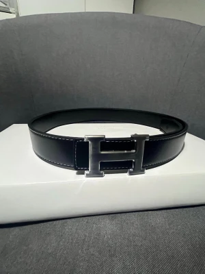 Svart hermm”Es bälte med silver logga - Säljer ett svart skärp med en rejäl metallspänne i form av ett H. Skärpet har vita sömmar längs kanterna och är gjort i slätt skinn. Perfekt accessoar för att lyfta vilken outfit som helst.
