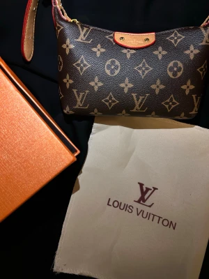 Louis Vuitton monogram axelväska - Säljer en ikonisk Louis Vuitton axelväska i klassiskt brunt monogrammönster med beige och röda detaljer. Väskan har guldfärgade metalldetaljer, justerbar axelrem och dragkedja. Insidan är fodrad med randigt tyg och har en liten ficka med LV-logga.
