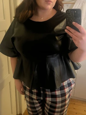 Svart peplumtop i skinnimitation - Säljer en svart peplumtop med rund hals och vida korta ärmar. Toppen har en snygg volang nertill och är gjord i glansig skinnimitation. Perfekt att styla med både jeans och kjol både till vardags men även till fest! Den är helt ny och aldrig använd! 