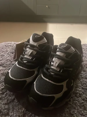 Svarta Asics Gel NYC - Säljer ett par svarta och vita Asics sneakers med coola detaljer och meshpartier för extra andningsförmåga. Säljer på grund av att de var för små för mig. Har aldrig används och är därför i helt nytt skick. Skorna har inga defekter alls. Skriv gärna till mig angående funderingar om skorna. 🤩👍