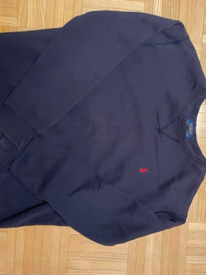 Mörkblå sweatshirt från Polo Ralph Lauren - Klassisk mörkblå sweatshirt från Polo Ralph Lauren i storlek S. Tröjan har rund halsringning, långa ärmar och den ikoniska röda broderade loggan på bröstet. Tillverkad i mjuk bomull för en skön och avslappnad look.