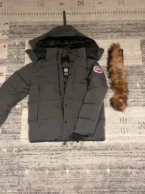 Mörkgrå Canada Goose dunjacka - Säljer en mörkgrå dunjacka från Canada Goose med avtagbar pälskant till huvan. Jackan har klassisk patch på ärmen, dragkedja och knappar framtill samt stora fickor. Perfekt för kalla dagar och riktigt snygg streetstil. Perfekt nu till vintern då det är kallt. Har haft på mig den en gång då den var lite för liten för mig! ( OBS… kan tänka mig gå ner nån hundra lapp vid snabbaffär.) Mvh