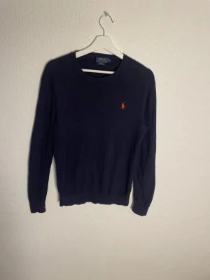 Mörkblå tröja från Polo Ralph Lauren - Klassisk mörkblå tröja från Polo Ralph Lauren i slim fit med rund hals och den ikoniska broderade loggan i orange på bröstet. Tillverkad i mjuk Pima bomull som känns riktigt skön mot huden. Perfekt till jeans eller chinos för en clean look.