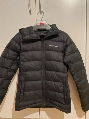 Svart dunjacka Peak Performance - Säljer en svart dunjacka från Peak Performance med huva och dragkedja framtill. Jackan har en quiltad design och logga broderad på bröstet samt ärmen. Perfekt för kalla dagar och riktigt snygg till alla outfits.