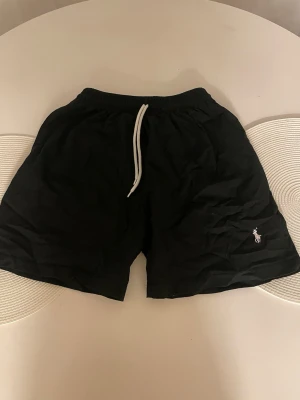Svarta shorts från Polo Ralph Lauren - Svarta shorts från Polo Ralph Lauren med vit snörning i midjan och klassisk broderad logga på ena benet. De har en avslappnad passform och är perfekta för sommaren. Materialet känns lätt och bekvämt.