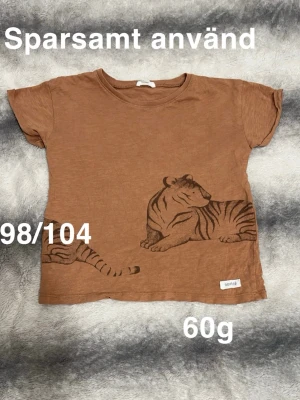 Brun t-shirt med tigertryck Newbie - Säljer en brun t-shirt från Newbie med ett coolt tigertryck på framsidan. T-shirten har korta ärmar, perfekt för en avslappnad stil. Passar dig som gillar djurmotiv och jordnära färger.🌸 Sparsamt använd. Storlek 98/104