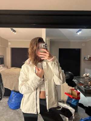 Vit vindjacka från Henri Lloyd - Snygg vit vindjacka från Henri Lloyd med huva och dragkedja framtill. Jackan har diskreta logodetaljer på ärmen och vid dragkedjan, samt praktiska fickor med knappstängning. Perfekt för blåsiga dagar och enkel att matcha med olika outfits.