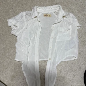 Vit croppad blus från Hollister XS - Säljer en vit croppad blus från Hollister i storlek XS. Blusen har korta ärmar, spetsdetaljer på axlarna och bröstficka. Knappar framtill och klassisk krage. Perfekt till sommaren för en fräsch och luftig look.