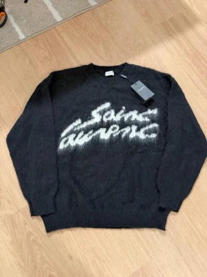 Svart Saint Laurent stickad tröja - Säljer en svart stickad tröja från Saint Laurent med vit fuzzy logga framtill. Tröjan har rund halsringning, långa ärmar och en lite lurvig känsla. Perfekt för dig som vill ha en statement piece med lyxig vibe.