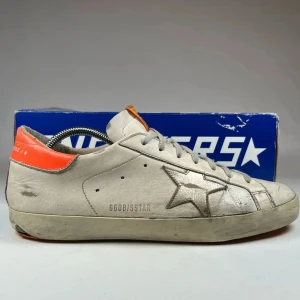 Golden gode superstar sneakers - Säljer ett par GGDB Superstar sneakers i vitt skinn med metallicstjärna på sidan och orange detalj vid hälen. Skorna har grå snörning, rund tå och klassisk platt sula. Tungan har GGDB-logga i orange. Perfekta för dig som gillar streetstyle och unika detaljer.