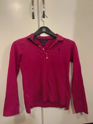 Mörkrosa henley från Ralph Lauren - Säljer en mörkrosa henley från Ralph Lauren med klassisk krage och tre knappar framtill. Skjortan har lång ärm och är tillverkad i mjuk bomull. Ikoniska loggan broderad på bröstet i mörkblått. Perfekt till en avslappnad men ändå stilsäker look för 12-14 åringa tjejer!!